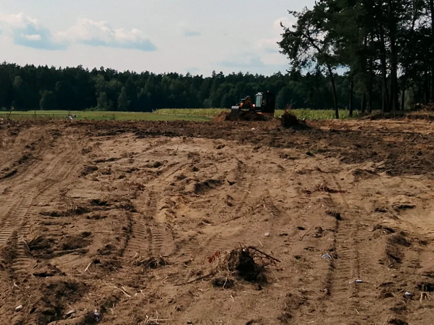 Wyrównany plac po niwelacji terenu ze śladami gąsienic koparki, w tle linia lasu